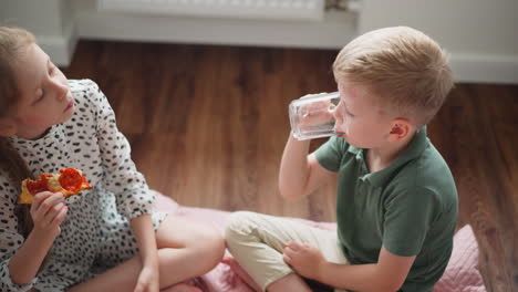 overhead view of little kids sitting on wooden floor having good time together indoors, boy drinking water from clear glass while girl holds slice of pepperoni pizza