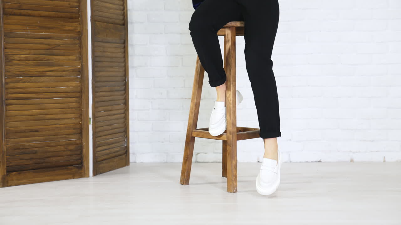 Girl model climbs up on the high wooden stool and puts her shoes on the footrest. White new modern footwear demonstration. White wall background.