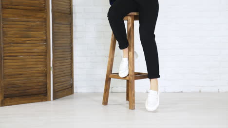 girl model climbs up on the high wooden stool and puts her shoes on the footrest. white new modern footwear demonstration. white wall background.