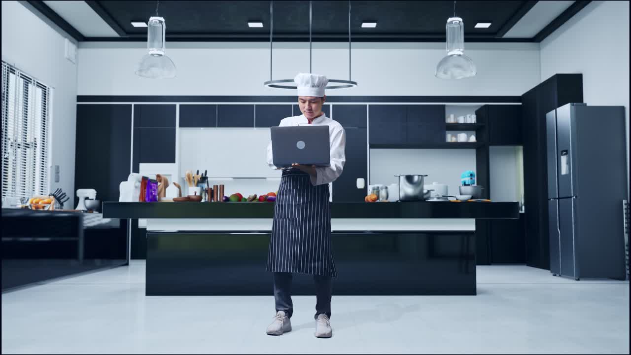 Chef asiatico a figura intera che utilizza un laptop, in piedi nella cucina di casa.
