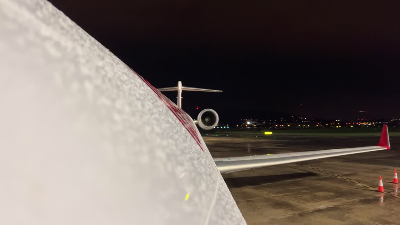 Vista della fusoliera superiore e dell'ala sinistra di un jet moderno dopo un acquazzone, in sosta all'aeroporto, mentre altri aerei stanno decollando sullo sfondo.