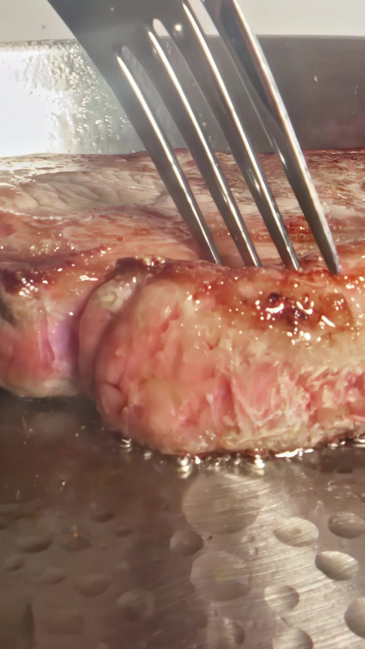 Una forchetta affonda in un spesso taglio di manzo marmorizzato su una piastra calda mentre la telecamera si avvicina, catturando il grasso che si scioglie, l'olio che bolle e l'interno rosato al culmine della rosolatura