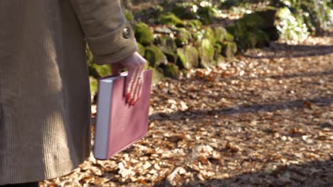 unrecognizable woman holds book in hand while walking in woods