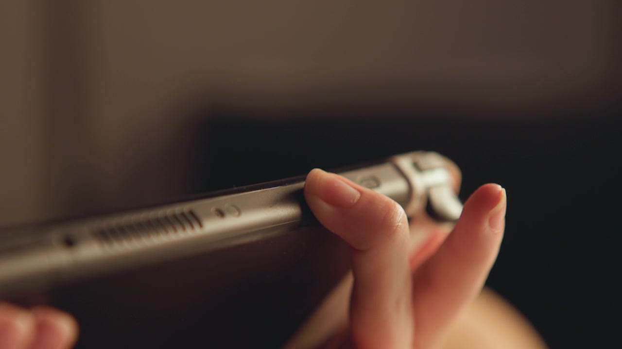 Primo piano ravvicinato mostra mani che tengono uno smartphone al bordo superiore mentre il pollice preme il pulsante laterale, con luce calda interna, messa a fuoco ridotta, sfondo sfocato e riflessi morbidi su superficie lucida