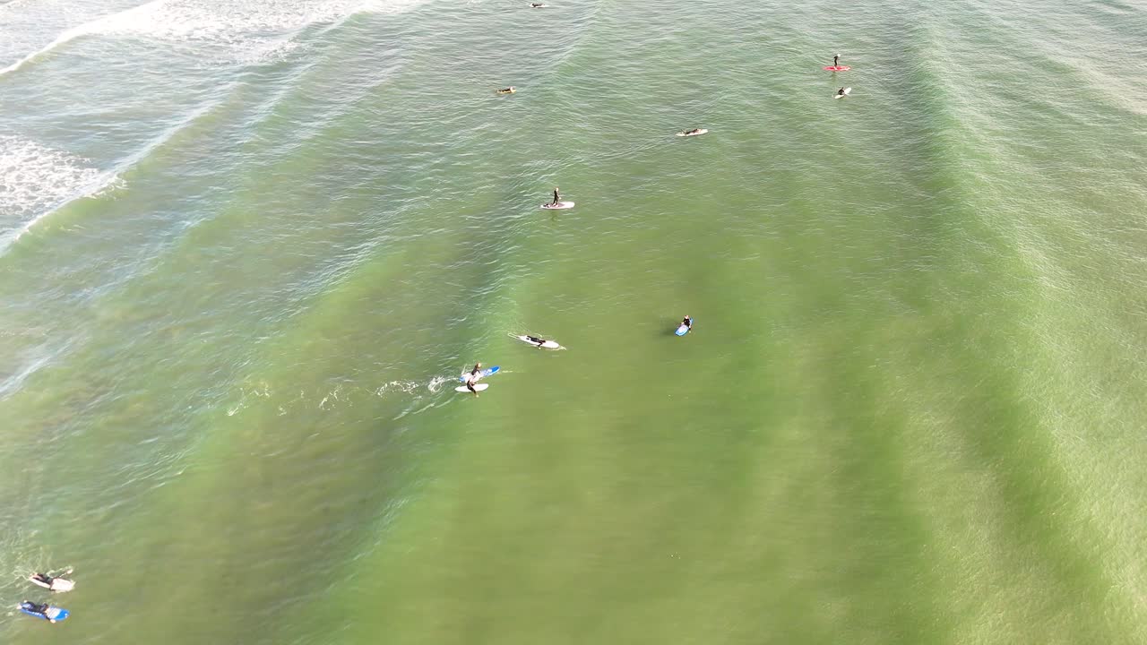 Aerial view of surfers paddling through shallow green waves at West Sands Beach, St Andrews, under bright daylight with steady overhead camera movement