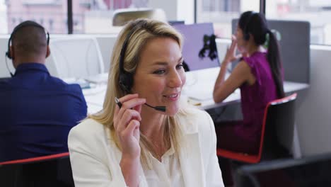 Caucasian-businesswoman-sitting-using-computer-talking-on-phone-headset-in-office