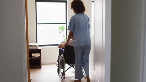 Mixed-race-female-doctor-assisting-senior-man-in-wheelchair-at-home