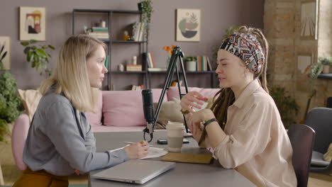 side view of two women recording a podcast talking into a microphone sitting at desk with laptop and documents 1