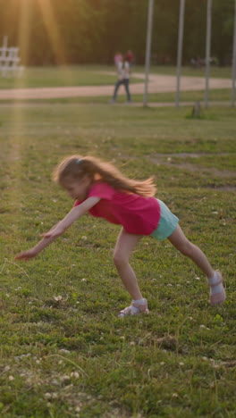 sporty little girl with ponytails does cartwheel on playground grass at sunset slow motion. gymnastic exercise in park. physical training of strong child