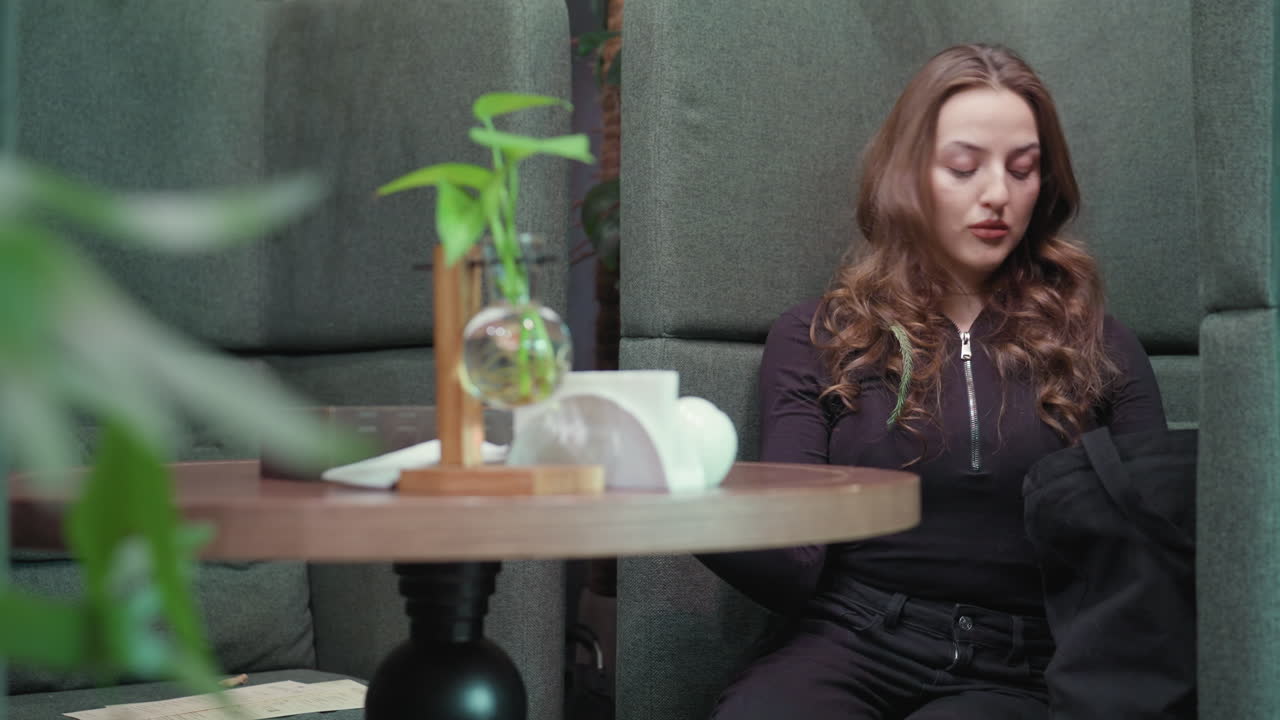 Blonde woman in black outfit sits inside cozy green booth reaching into black bag to bring out books while soft indoor lighting, minimal tabletop decoration, and surrounding plants set calm atmosphere