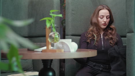 blonde woman in black outfit sits inside cozy green booth reaching into black bag to bring out books while soft indoor lighting, minimal tabletop decoration, and surrounding plants set calm atmosphere
