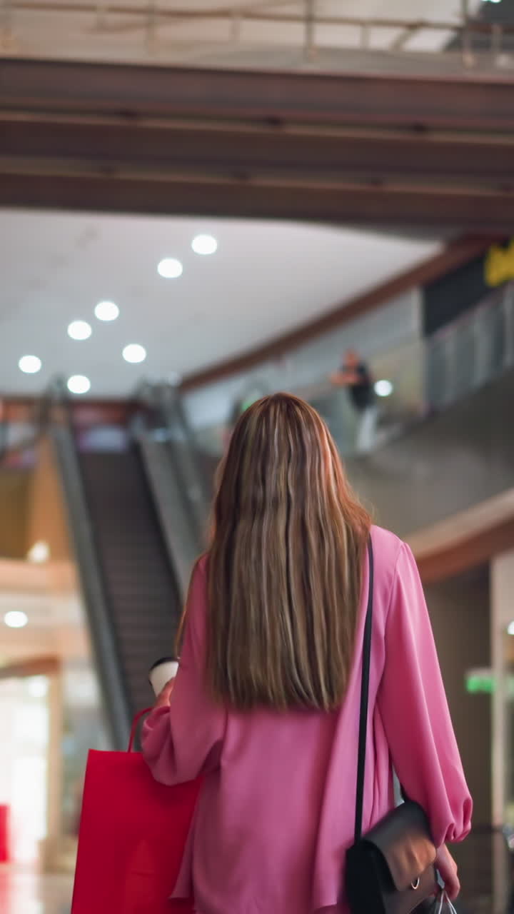Back view of woman in pink dress holding coffee cup and walking through brightly lit mall, blurred background features stores, lights, and bokeh effect, while people walk in the distance