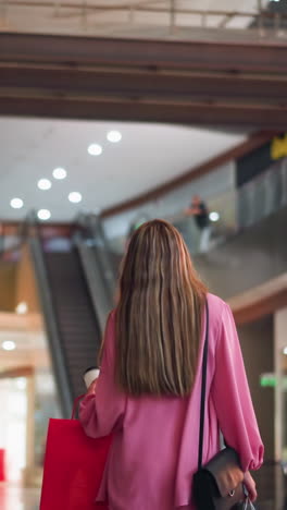 vista trasera de una mujer con un vestido rosa sosteniendo una taza de café y caminando a través de un centro comercial brillantemente iluminado, el fondo borroso presenta tiendas, luces y efecto bokeh, mientras la gente camina en la distancia