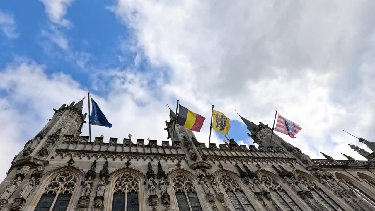 Low-angle view of colorful flags waving atop ornate Gothic city hall in Bruges, Belgium