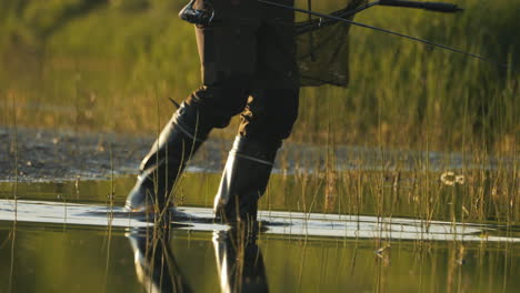 Pescador-Con-Botas-De-Goma-En-Medio-De-Aguas-Poco-Profundas-Y-Juncos-Al-Atardecer,-Tranquilidad-Otoñal