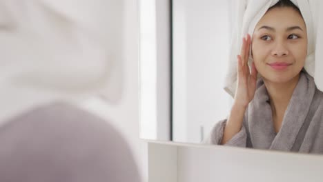 Video-of-portrait-of-smiling-biracial-woman-with-towel-on-hair-looking-in-mirror-in-bathroom