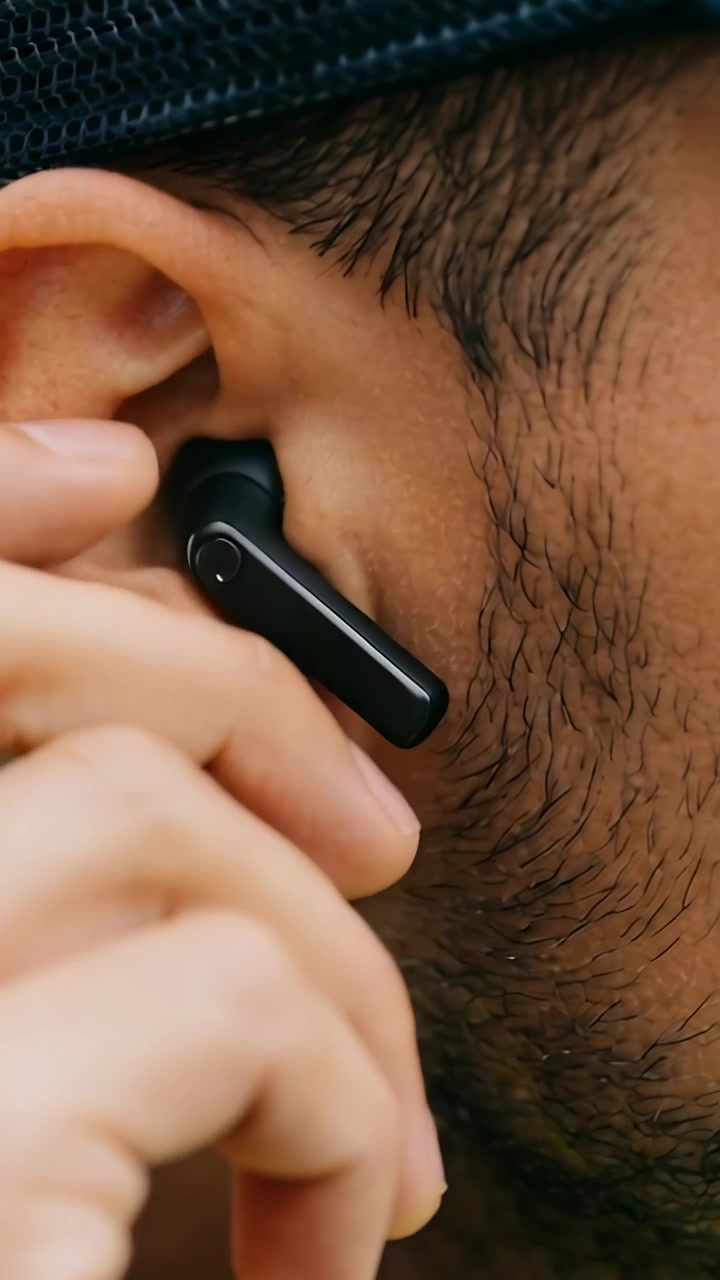 Video verticale: Mano che raggiunge un uomo con un berretto che sistema un auricolare nero per farlo aderire, in primo piano con un punto giallo.