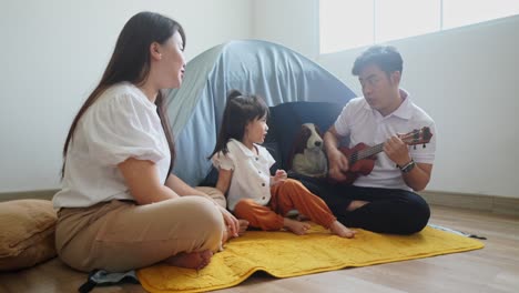 행복한 아시아 어린 소녀와 어머니는 아버지가 집에서 텐트와 함께 우쿨레를 연주하는 동안 노래합니다.