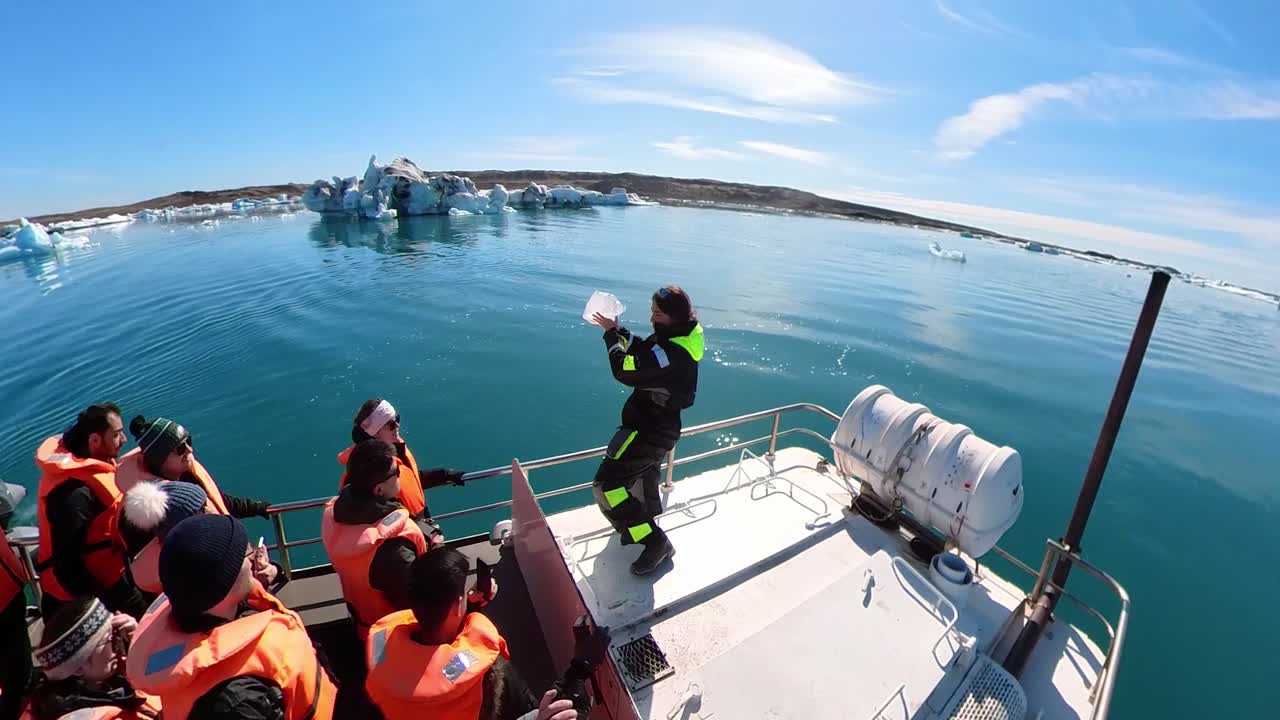 A young woman holding a piece of ice, shares its beauty with her companions on the boat, against the tranquil backdrop of a calm, icy winter ocean under a serene blue sky.