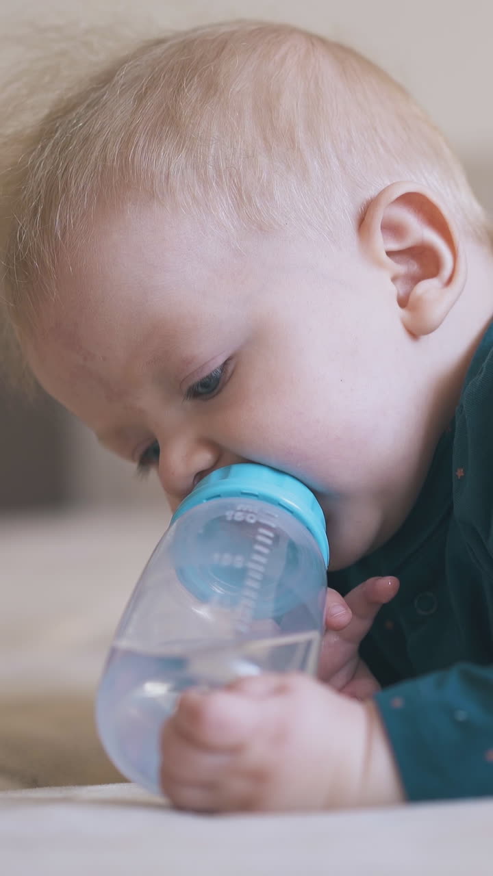 adorable blond baby boy nibbles teat of bottle with water lying on large soft bed in spacious children room close view