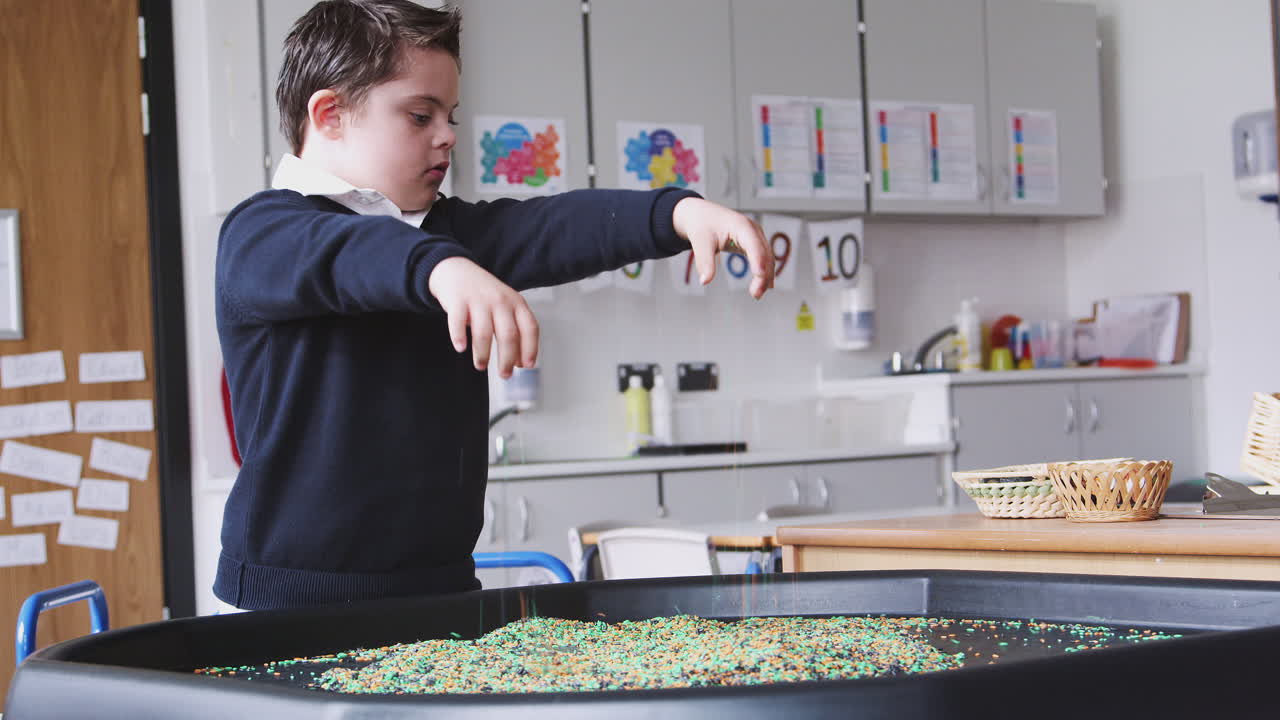 Primary schoolboy with Down Syndrome stands using a sensory play tub in a classroom, side view
