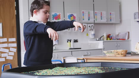 Primary-schoolboy-with-Down-Syndrome-stands-using-a-sensory-play-tub-in-a-classroom,-side-view