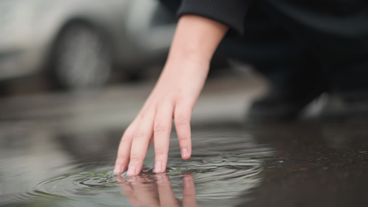 Caucasian woman hand touching puddle, urban street reflection and gentle ripple rings, parked cars blurred in background, rainy asphalt, quiet reflective mood and tactile curiosity