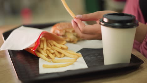 woman s hand reaching for fries on a tray with a partially eaten burger and coffee cup, after picking the fry, she takes a bite and drinks from her coffee, with soft bokeh lighting in the background