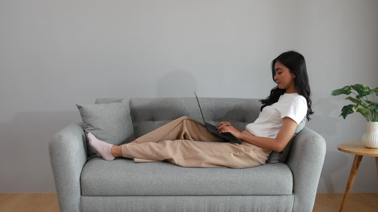 Una giovane donna asiatica si rilassa comodamente a casa, concentrata sul suo laptop con un sorriso rilassato.