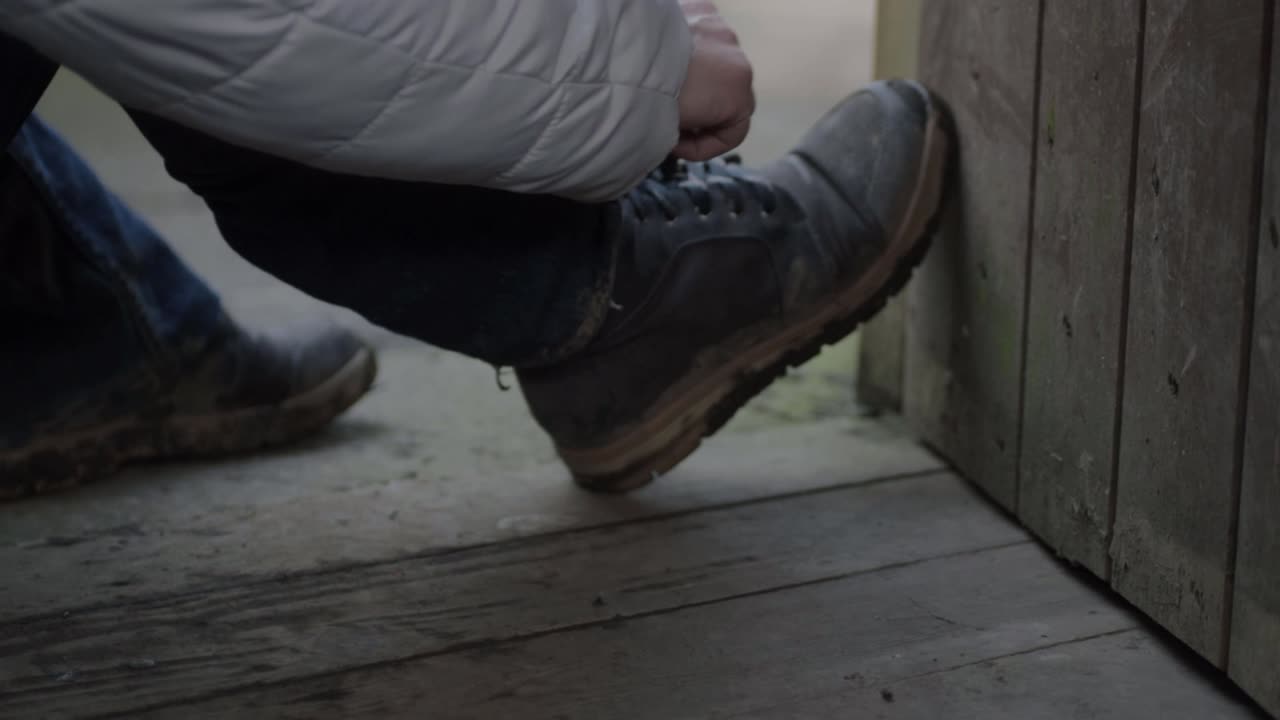 Hiker stops in rustic wooden building doorway to tie shoelaces on walking boots wide shot