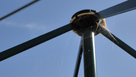 Pan-around-large-cob-spider-web-on-green-clothes-line-gently-blowing-in-breeze-with-bugs-against-blue-sky-background