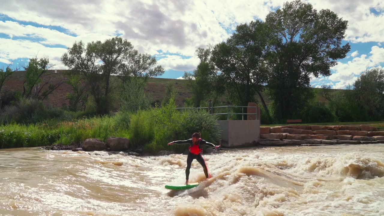 Joella surffaajat surffaavat paikallaan pysyvällä aallolla Montrosen vesiurheilupuistossa Montrosen kaupungissa Coloradossa, Uncompahgre-joella aurinkoisena iltapäivänä. Maisemaan kuuluvat Cerise Park ja McNeil Fields, joella on suuri virtaus lumen sulamisen ja mutaisen sedimentin vuoksi.
