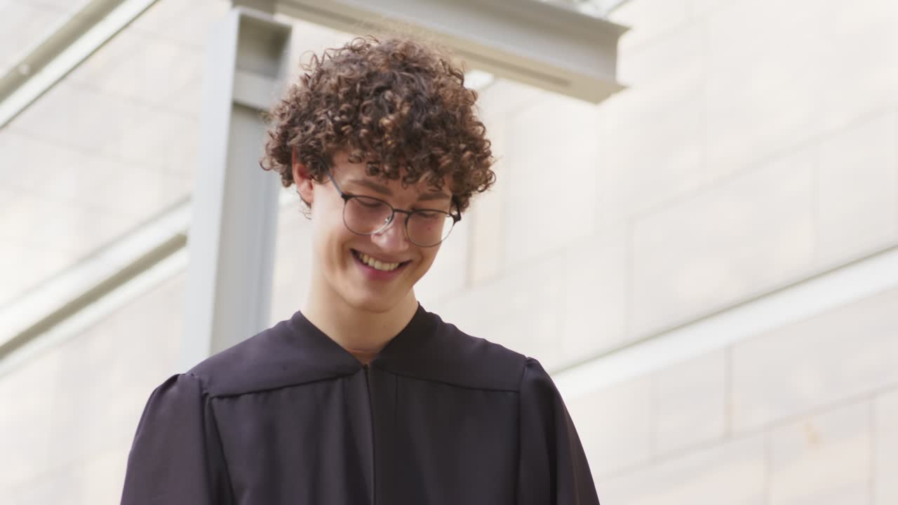Man wearing black graduation gown reading smartphone outside building, smiling, chuckling, nodding