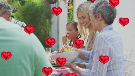 animación de globos de corazón sobre una feliz familia caucásica comiendo juntos en el jardín