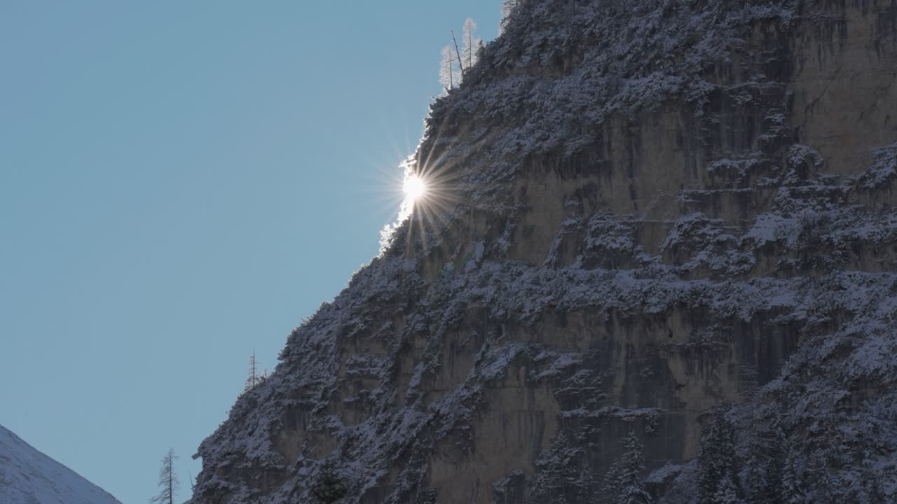 Bright winter sun slowly hides behind the rugged cliff covered with snow-dusted pine trees. Parallax video.