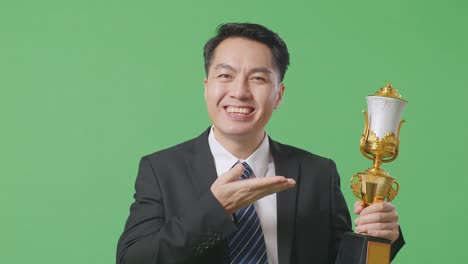 close up of asian business man in a suit and tie with a gold medal pointing to a gold trophy in his hand and smiling being proud winning as the first winner on green screen background in the studio