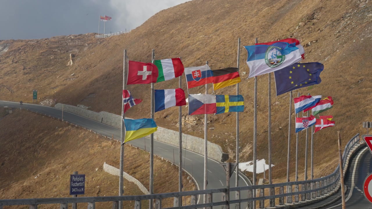 Colorful flags of various countries stand tall along a winding mountain pass, fluttering gently in the wind. The landscape is rugged, with a cloudy sky above, suggesting varying weather conditions.
