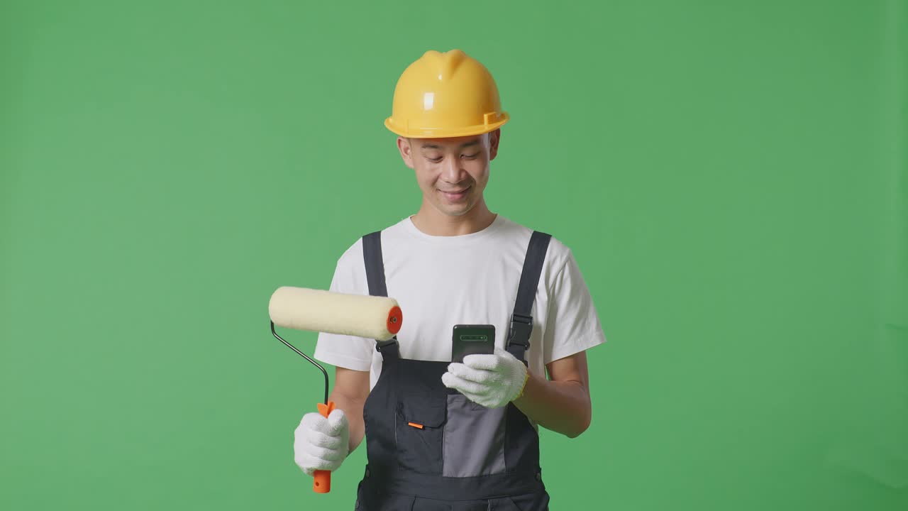 Asian Man Painter Wearing Safety Helmet Smiling And Using Smartphone While Standing In The Green Screen Background Studio