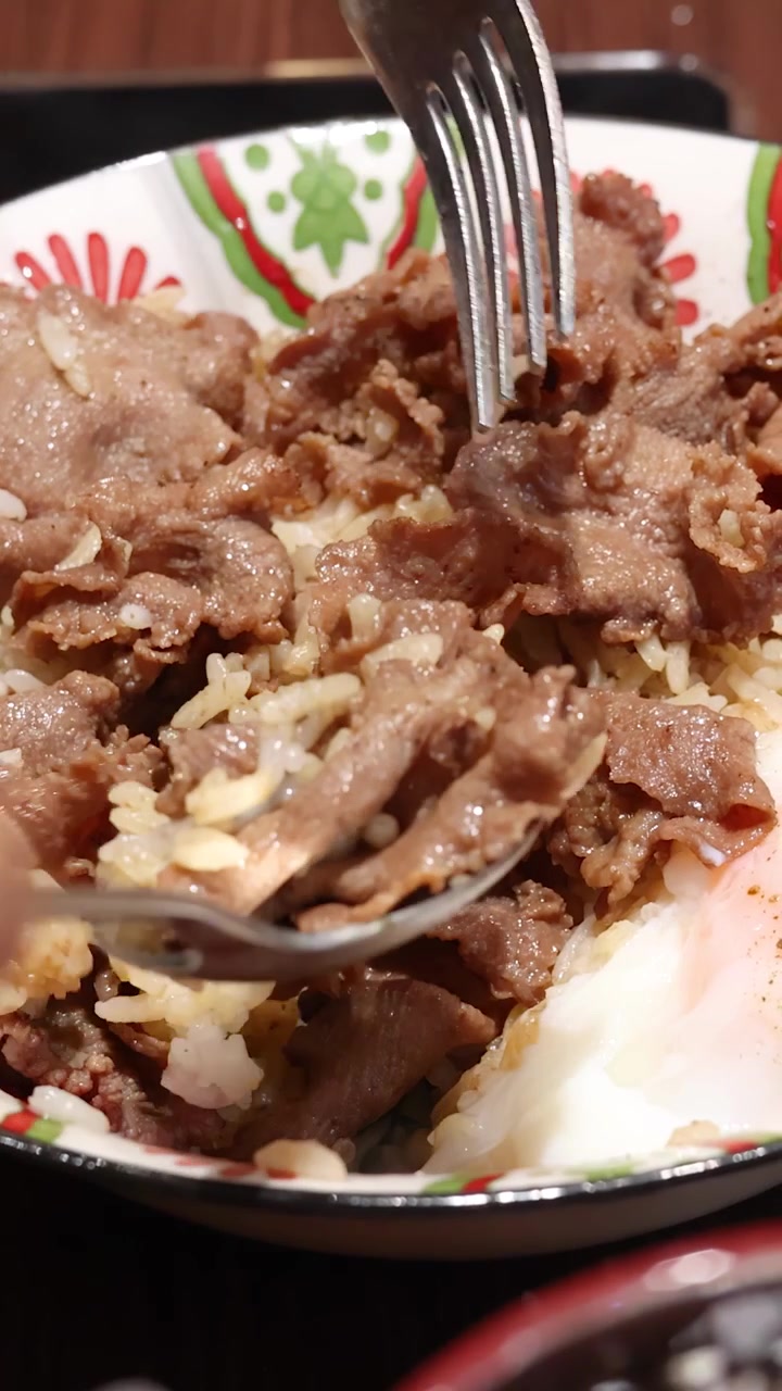 Primer plano de manos usando una cuchara y un tenedor para mezclar carne de res en rodajas, arroz y un huevo pasado por agua en un tazón estampado bajo una cálida iluminación interior.