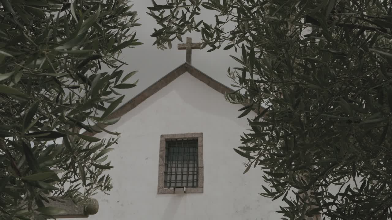올리브 나무 가지에 둘러싸여 있고 꼭대기에 십자가가 있는 작은 흰색 교회의 정면 모습