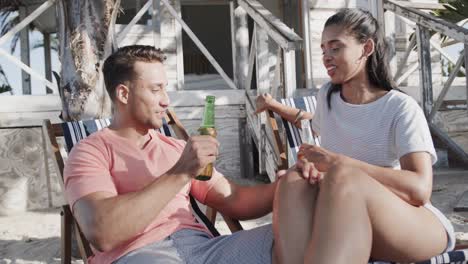 happy diverse couple sitting and drinking a toast with beers outside beach house, in slow motion