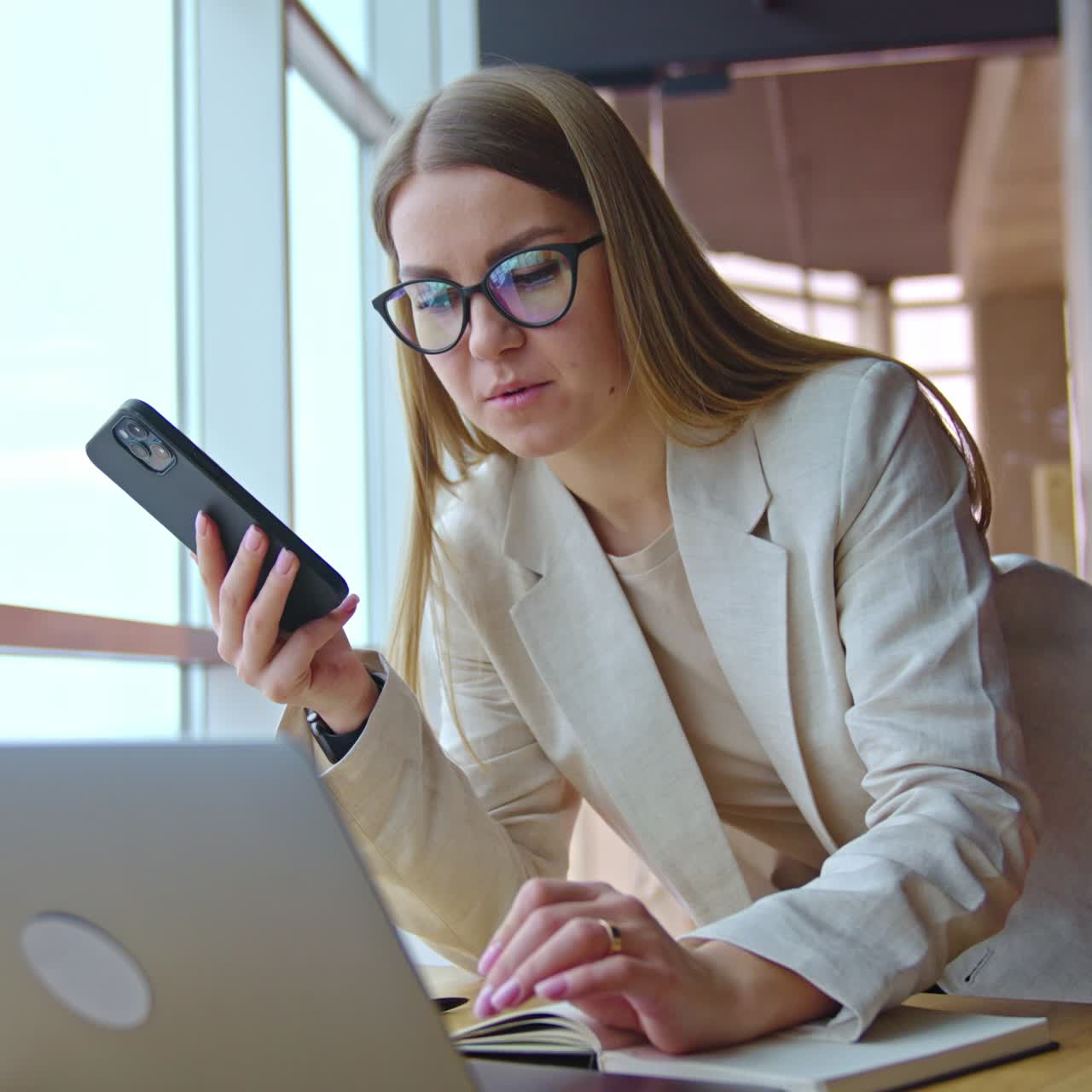 Blonde young woman speaking on the phone leaning on the table. Female employee talks on the phone and works on laptop. Office at backdrop