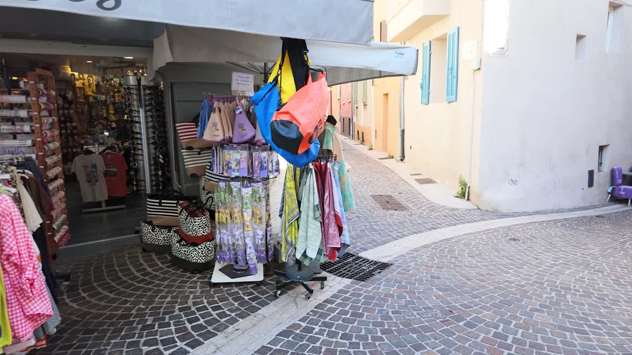 Uma câmera faz um movimento panorâmico de uma vitrine de boutique, atravessando uma rua estreita de paralelepípedos sob a luz do sol forte.
