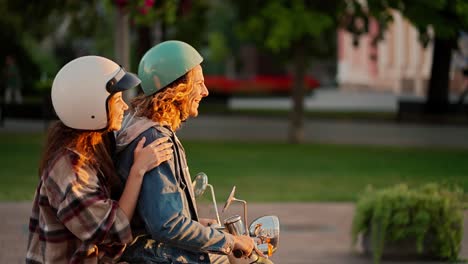 Side-view:-a-guy-with-long-curly-hair-in-a-green-moped-helmet-and-a-denim-jacket-rides-with-his-brunette-girlfriend-in-a-plaid-shirt-on-a-moped-along-the-street-at-a-golden-sunset-in-a-summer-city