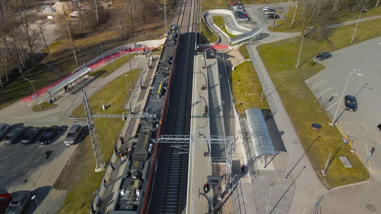Aerial drone footage flying along backwards with an electric orange colored train coming to train station in Laagri Estonia during a spring or autumn sunny day while leafless trees and grass is around