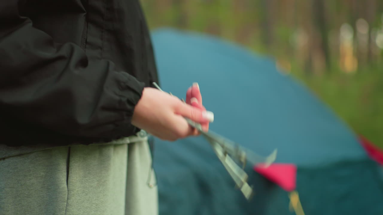 Close up of lady holding and twirling metal pegs outdoors with forest camping tent in background, wearing casual outfit, preparing for camping setup in nature with blurred greenery and gear in view