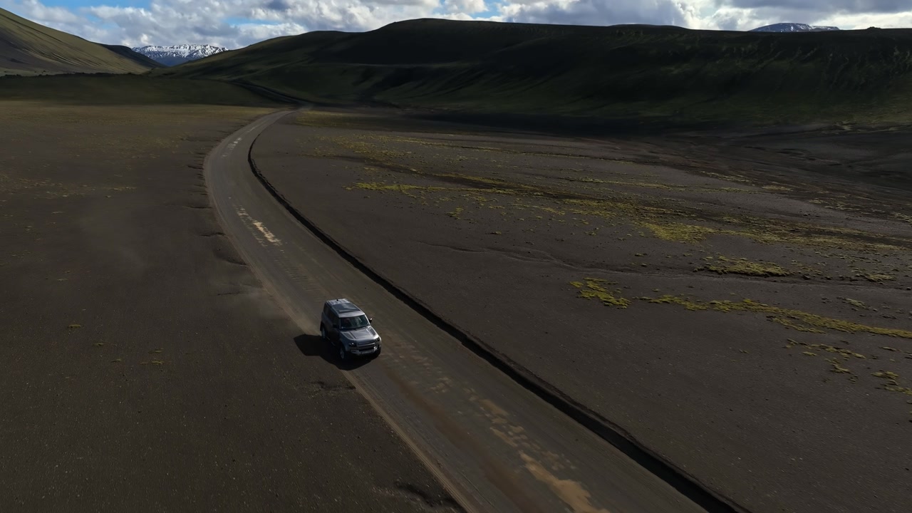 Drone aerial of a lone 4x4 SUV driving a winding dirt road across Iceland’s black volcanic highlands, with dramatic shadows and wide copy space.