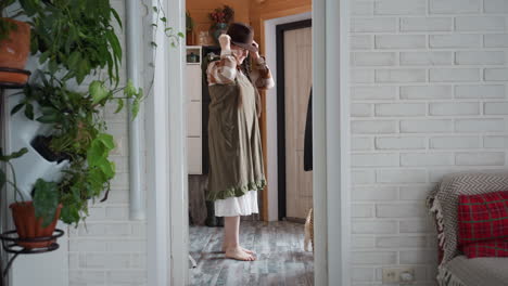 inside view from door shows woman in olive green gown over striped shirt walking barefoot on wooden floor toward corner to pick hat, with green plant outside and dog inside wagging tail