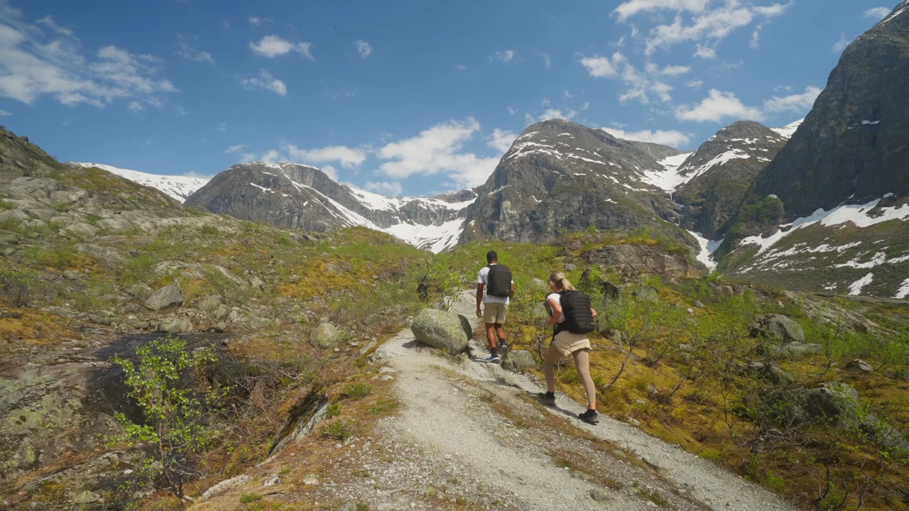 Inquadratura statica di una coppia di escursionisti che sale un sentiero di montagna in Norvegia, con cime innevate e un cielo azzurro limpido. Un suggestivo paesaggio alpino che cattura l'avventura, il viaggio e lo stile di vita all'aria aperta.