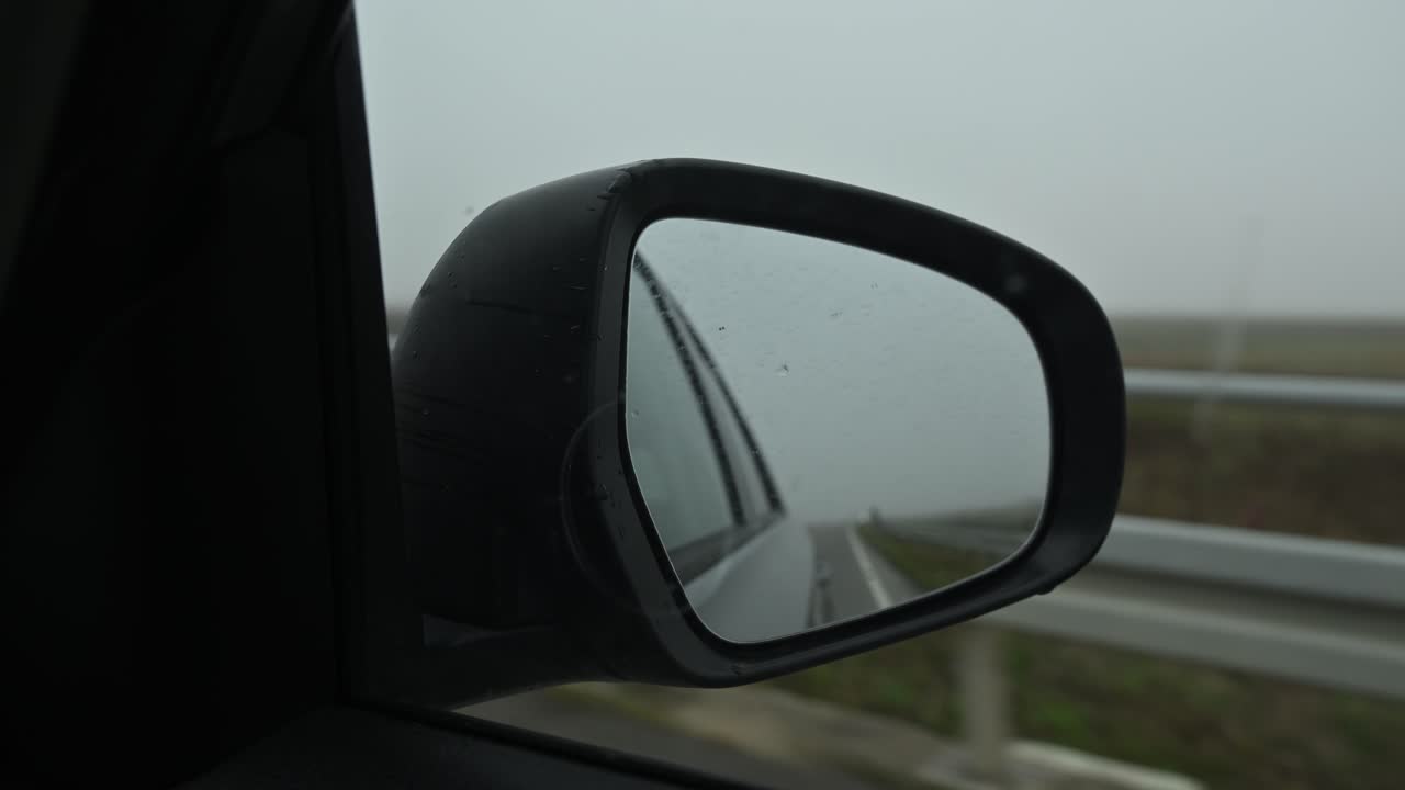 Car side mirror reflecting a foggy autumn countryside scene while driving along a rural road. Selective focus